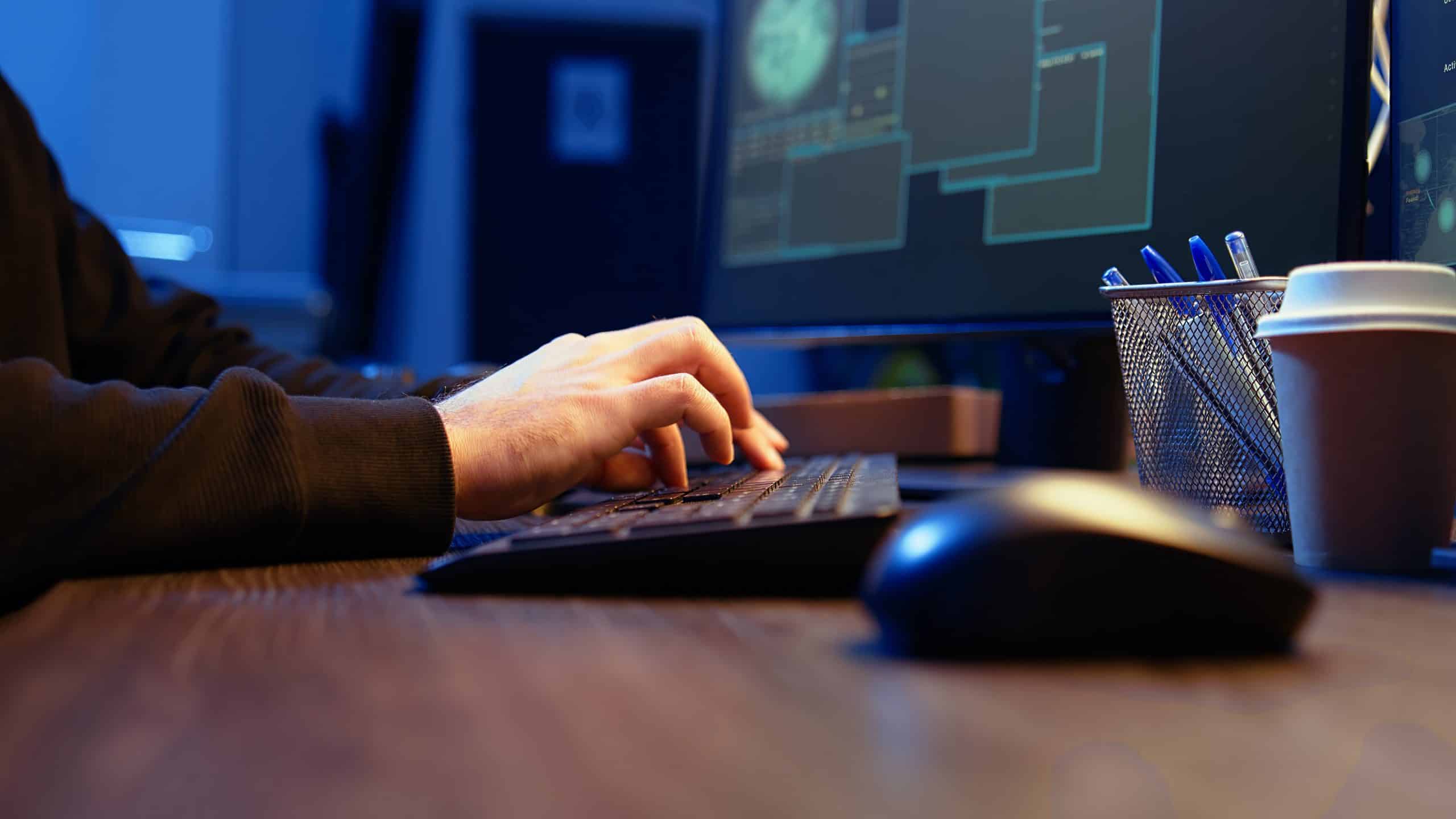 Manos escribiendo en un teclado en una oficina oscura con luz azul.